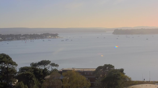 A coastal scene showing a calm body of water with several small boats scattered throughout. In the foreground, there are some trees and buildings, while in the background, a waterfront with houses lines the shore. The sky is clear with soft pastel colors, indicating either dawn or dusk. The water reflects the light, creating a tranquil atmosphere.