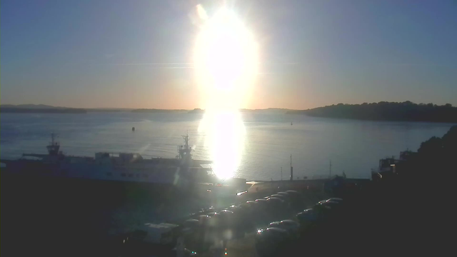 A scenic view of a harbor at sunset, with the sun low in the sky, reflecting brightly on the water's surface. A large boat is docked at the left side of the image, partially obscured by glare from the sunlight. In the foreground, there is a parking area filled with cars, and the background features a distant shoreline with trees silhouetted against the colorful sky. The horizon is slightly undulating, suggesting hills in the distance. The overall mood of the image is calm and serene.