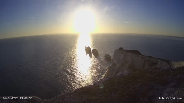 A scenic view of the ocean during sunset, with the sun shining brightly in the sky, reflecting on the water. In the foreground, there are cliffs with a rocky outcrop in the water. The colors of the sky transition from blue to warm hues of orange and yellow. The landscape appears tranquil, and a small structure is visible on the cliff. The date and time are displayed in the bottom left corner.