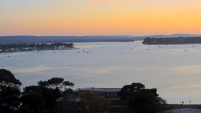 A tranquil view of a calm body of water during sunset, reflecting soft hues of orange and pink in the sky. Small boats are scattered across the water, with a tree line and distant hills visible in the background. In the foreground, there are dark silhouettes of trees and a low building with windows, partially obscured by foliage.
