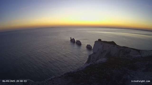 A panoramic view of a coastline at sunset. The sky transitions from blue to orange and purple hues, reflecting soft light over a calm sea. Prominent white cliffs are in the foreground, with three distinct rock formations emerging from the water, to the left of the image. A small building is perched atop a cliff on the right side, with a faint outline of land visible in the distance. The scene conveys a tranquil and picturesque moment by the sea.