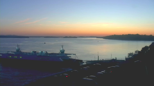 A serene waterfront scene at sunset, featuring calm waters reflecting shades of orange, pink, and blue in the sky. On the left, a large ferry boat is docked with bright lights illuminated on its deck. The background includes distant hills and a horizon line where the water meets the sky. Silhouetted cars are parked on the right side of the image, creating a contrast against the evening light.