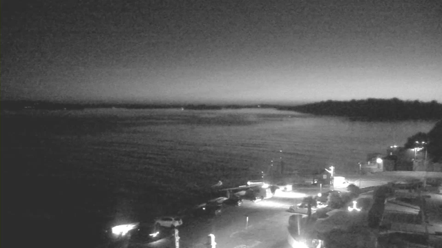 A black and white image of a calm waterfront scene at dusk. The water reflects soft, subtle waves and has a slight mist across it. In the foreground, there is a dimly lit pier and several boats docked. A few lights illuminate the pier and surrounding area, while the land in the background includes trees and a distant shoreline, slightly obscured by the fading light of the evening sky.