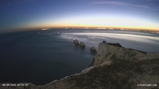 A coastal scene at dusk featuring rocky cliffs on the right side and a calm sea reflecting the fading light. In the distance, faint lights from a town can be seen along the horizon. The sky transitions from deep blue at the top to warm orange near the horizon. On the left side, two rock formations jut out of the water, with a light on one of them. The foreground shows uneven terrain with patches of grass and stones. The timestamp indicates it is about 20:59 on April 6, 2025.