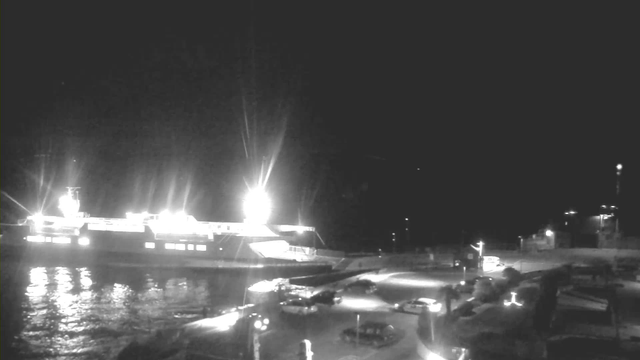 A nighttime scene at a harbor with a brightly lit ferry docked at the left side of the image. The water reflects the lights of the ferry and other illuminated areas. In the foreground, there are several parked cars, along with dim streetlights and a winding path leading towards the water. The sky is mostly dark with a few faint light sources visible.