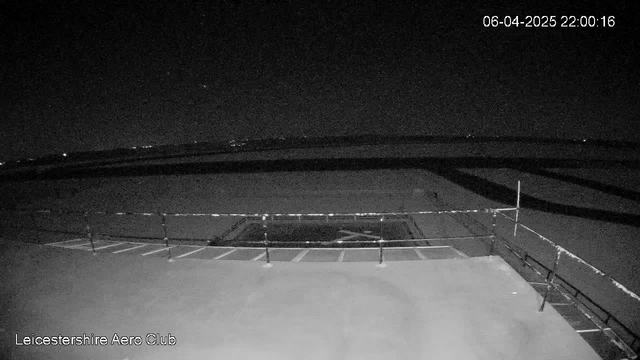 A dark scene depicts a flat rooftop with a railing on the left side. In the background, distant lights are faintly visible against the night sky. There is a large, dark area in the center, possibly a landing space or empty lot. The date and time are stamped in the upper right corner of the image.