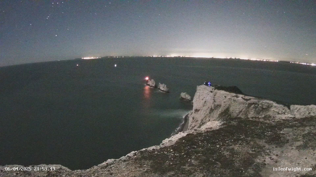 A night scene showing a rocky cliff overlooking the sea. In the foreground, there are jagged white cliffs with some vegetation. In the distance, several small lights from boats are visible on the water. A lighthouse emits a red light on one of the rocky formations in the sea. The sky is dark, sprinkled with stars, and there are faint lights from a distant shore on the horizon. The overall atmosphere is calm and serene.