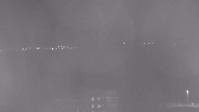 A dark nighttime scene showing faint lights in the distance, likely from buildings or streetlights, against a hazy, indistinct background. Some light reflections are visible, but the overall visibility is low, creating a muted atmosphere.