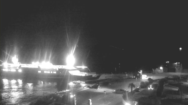 A dimly lit waterfront scene at night features a large ferry boat docked at a terminal. The ferry is illuminated by bright lights, reflecting on the dark water. In the foreground, there are a few parked cars near the terminal, and an empty parking area is visible. The surroundings are mostly dark, with minimal lighting from street lamps and the ferry itself.