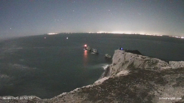 A nighttime view of a coastal scene featuring cliffs on the right side and a calm sea below. Several rocks jut out from the water, with one having a red light on it. The background shows city lights along the horizon, and a few stars are visible in the sky. The overall ambiance is serene, with a dark blue sky and illuminated areas reflecting off the water.
