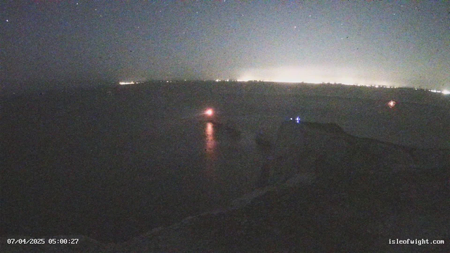 A night seascape showing a dark shoreline with a faint glow from distant city lights along the horizon. A small boat with a bright red light is visible on the water, reflecting on the surface. The scene is illuminated by stars in a dark sky, and the date and time are displayed at the bottom of the image.