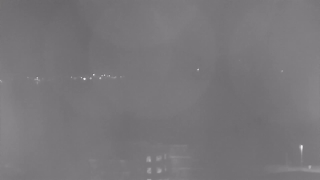 A dimly lit nighttime scene captured by a webcam. In the background, several small, indistinct lights are visible, scattered across a dark landscape, indicating distant buildings or streetlights. In the foreground, there appears to be a structure with faint outlines and a few lit windows, suggesting a residential or commercial building. The image has a grainy appearance, with a hazy quality obscuring details.