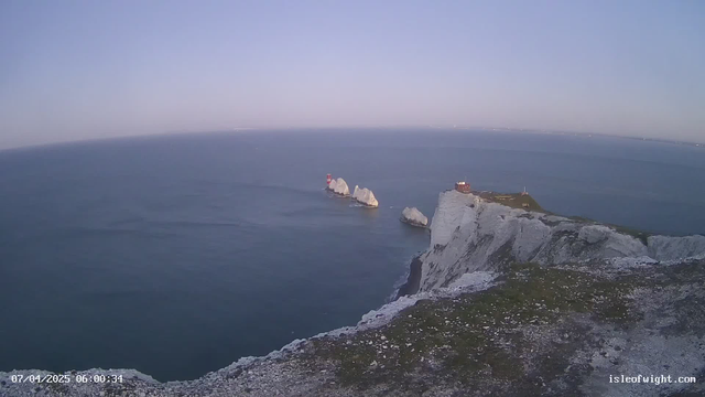 A coastal scene at dawn featuring rocky cliffs on the right side, with waves gently lapping against the shoreline. Three large rock formations are visible in the water, with two red lighthouses positioned on them. In the background, a smooth horizon meets a clear sky tinged with soft blue. The ground in the foreground is covered with grass and rocky debris, suggesting a natural, rugged environment.