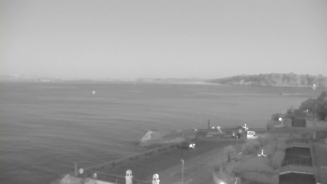 A black and white image of a waterfront scene. The foreground shows a paved area with a slight incline leading towards the water. There are several light posts and a few vehicles parked near a building on the right. The background features calm water extending to the horizon, with a small boat visible. In the distance, hills can be seen, partially shrouded in mist. The sky is gray, suggesting overcast weather.