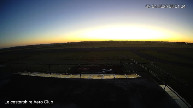 A wide view of a landscape at dawn. The sky transitions from deep blue at the top to a warm orange on the horizon, indicating sunrise. In the foreground, there is a flat rooftop with a railing, leading to a grassy area below. A runway is visible in the distance, bordered by a fence. The overall scene is peaceful and represents an early morning at an outdoor location.
