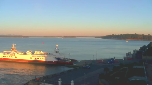 A calm body of water is visible with a large white and red ferry docked near a waterfront. The sky transitions from light blue to soft orange, indicating either dawn or dusk. In the background, there are distant trees lining the shore, and small boats can be seen on the water. The foreground features a paved area with a few parked cars and structures. There is a buoy marked by a red sphere floating in the water.