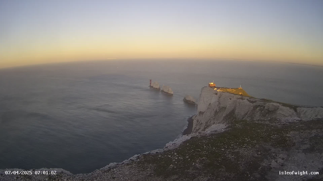 A coastal view at dawn showing cliffs on the right with a small building illuminated by the early light. The sea is calm and reflects the soft colors of the sky, ranging from pale blue to golden hues. There are rocky formations emerging from the water, with a red lighthouse standing out prominently in the distance. The foreground features grassy areas at the cliff's edge.
