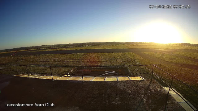 A clear sky at sunrise with a bright sun in the background. In the foreground, a grassy area is visible, accompanied by a flat runway leading to the horizon. A simple fence borders the left side, and a small white cross marking is present on the ground. The overall scene conveys a calm morning atmosphere at an aerodrome or airfield.