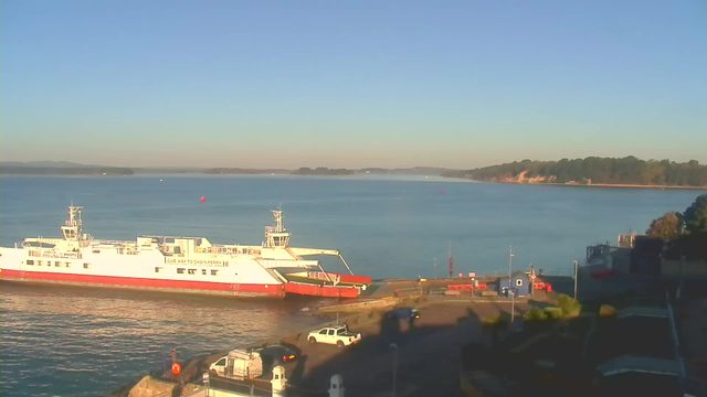 A ferry boat is docked at a pier on a calm body of water. The sky is clear and blue, and the water reflects the sunlight. In the distance, there are trees lining the shore. Near the dock, there are a few vehicles parked, and a red buoy is visible in the water nearby. The scene is tranquil, with no visible people.