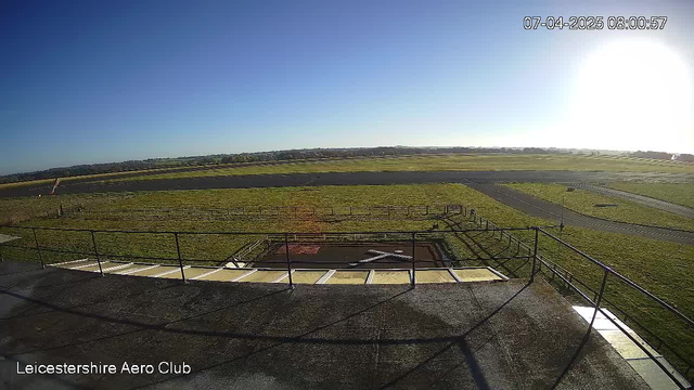 A clear blue sky fills the upper portion of the image, with bright sunlight streaming from the right-hand side. Below, a grassy area stretches out, leading to a runway that extends into the distance. In the foreground, a helipad with a large white cross is visible, surrounded by a railing. The scene suggests a tranquil morning at the Leicestershire Aero Club.