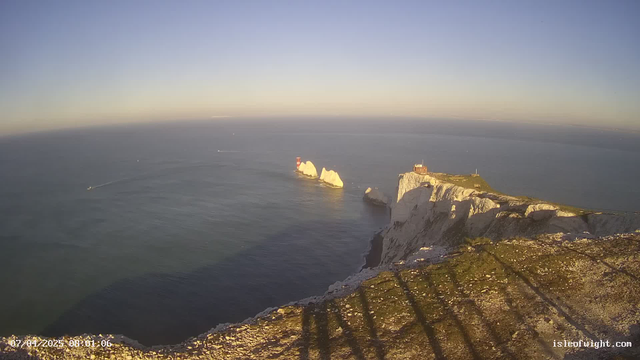 A coastal view at dawn featuring white chalk cliffs meeting the sea. In the distance, there are two small islands with red lighthouses. A boat is creating a ripple in the water as it travels across the smooth surface of the ocean. The sky transitions from blue to light shades near the horizon, indicating early morning. Shadows cast on the rocky foreground suggest the presence of features or structures above.