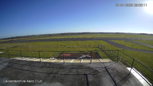 The image shows a clear blue sky above an expansive green field. In the foreground, there is a black and white “X” marking on the ground, likely indicating a helipad or landing area. Behind the marking, a fenced area is visible, with a long asphalt runway stretching across the midground. The scene is bathed in sunlight, creating a bright and open atmosphere.