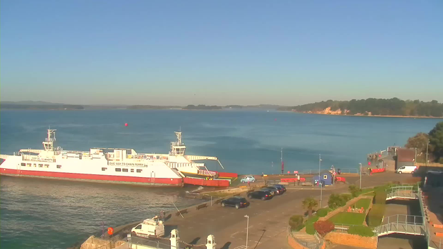 A view of a calm sea under a clear blue sky, with a large white and red ferry boat docked at a pier. Several vehicles are parked along a paved area near the waterfront. In the background, there are hills and trees lining the shore. A few people and structures can be seen near the dock.