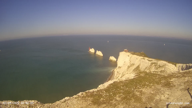 A panoramic view of the ocean and cliffs under a clear blue sky. The white chalk cliffs rise sharply from the water, with some rocky formations and a lighthouse visible on the left side. Small sailing boats dot the calm sea in the distance. The foreground features a grassy area at the cliff's edge, leading down towards the beach below. The overall atmosphere is tranquil and sunny.