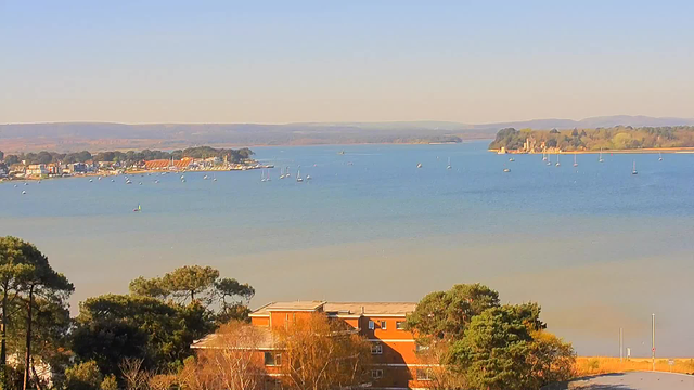A scenic view of a calm bay with multiple small boats anchored. In the foreground, there is a brick building partially hidden by trees. In the background, a shoreline with various structures can be seen alongside the water. The sky is clear and blue, with rolling hills visible in the distance. The color of the water transitions from blue to a lighter shade near the shore.
