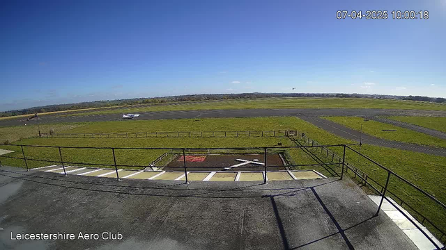 The image shows an aerial view from a webcam mounted on a structure at Leicestershire Aero Club. The scene features a wide expanse of green grass and a paved runway under a clear blue sky. In the foreground, there is a platform with a railing and white and yellow markings on the ground, including a large white cross in a designated area. In the distance, a small aircraft is seen on the runway, and there is a slight indication of more aircraft positioned further back on the field.