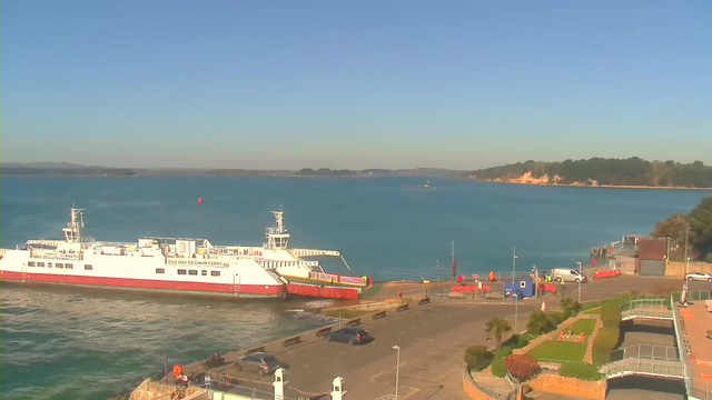 A bright, clear day at a waterfront area. In the foreground, a white and red ferry is docked at the pier, with its ramp extending onto the shore. Several cars are parked nearby on a roadway, and some people are seen walking. In the background, the water is calm and blue, with a few boats in the distance. On the right, there is a building near the water's edge, surrounded by greenery and flowers, and a sandy beach further along the shore. The horizon shows a tree-lined coastline under a clear sky.