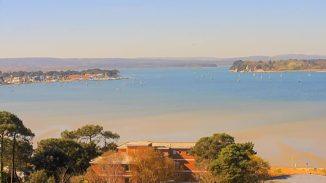 A scenic view of a calm waterway, with several small boats sailing on the water. In the background, a shoreline with trees and buildings is visible, extending along the water's edge. The sky is clear and blue, with gentle hills appearing in the distance. The foreground features greenery and a building with an orange exterior. The overall atmosphere conveys a serene and peaceful setting.