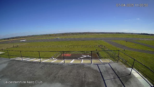 A clear blue sky with a few clouds is visible over a grassy airfield. In the foreground, there is a railing along a rooftop with a gray surface. Below the railing, a large red marked area with a white 'X' is present, indicating it might be a helipad or similar. To the left, a small airplane is taxiing on the runway, while more grassy fields and another airplane can be seen in the background, along with dirt taxiways. The overall scene conveys a sunny day at an airfield.