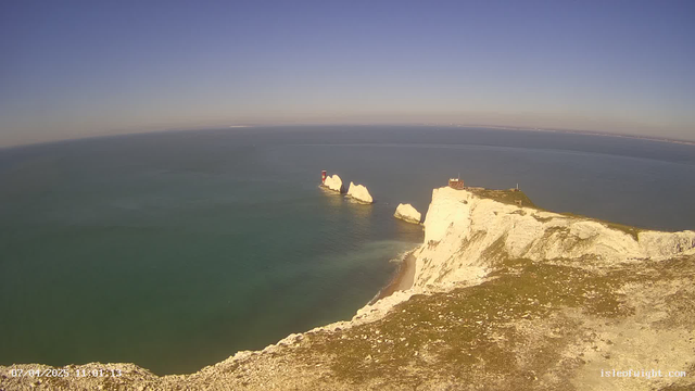 A panoramic view of a coastline featuring white cliffs on the right. The ocean is calm, with varying shades of blue and green. In the distance, several small islands are visible, and a red lighthouse stands on one of the rocky outcrops. The sky is mostly clear with a few wispy clouds. The foreground has a grassy area with some rocks.