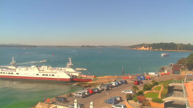 A scenic view of a harbor on a clear day. In the foreground, a large white ferry with red accents is docked, while a smaller boat is visible moving across the blue water. The waterfront is lined with a parking area filled with cars and people walking. On the right, there is a small building and greenery with pathways. In the background, the coastline features cliffs and trees under a blue sky.