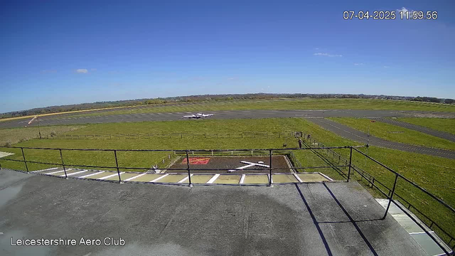 A wide view of an airfield under a clear blue sky. In the foreground, there is a low railing along a terrace, with a small grassy area featuring a red patch and a white cross shape marked on the ground. An aircraft is seen on the runway in the background, taking off or landing. The airfield is surrounded by green fields, and there are some trees and distant hills visible on the horizon. The time displayed is 11:59 on April 7, 2025.