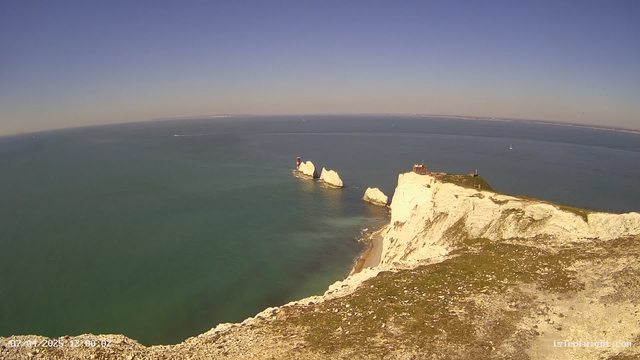 A coastal scene featuring white chalk cliffs on the right side of the image. In the foreground, there is grass and rocks. The ocean is visible with calm, blue waters extending to the horizon, where a few small boats can be spotted. On the cliffs, there are notable tall rock formations and a building near the shoreline. The sky is clear with a gradient from light blue near the horizon to a deeper blue above. The date and time are displayed in the bottom left corner.