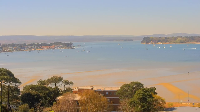 A scenic view of a calm water body, with boats scattered across the surface. In the foreground, there are trees and a building visible. The background features a shoreline lined with houses and greenery, transitioning into low hills under a clear blue sky. The water shows ripples and sandy patterns, indicating shallow areas.