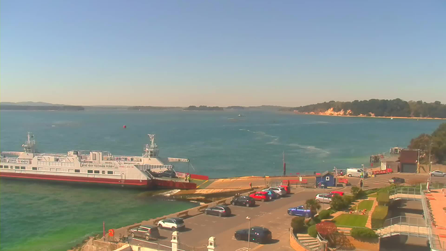 A ferry is docked at a pier, surrounded by a vibrant blue sea under a clear sky. In the foreground, several cars are parked in a lot, with a mix of colors including red, blue, and white. To the right, there are people walking and a blue structure near the water. The distant shoreline features green trees and sandy cliffs.
