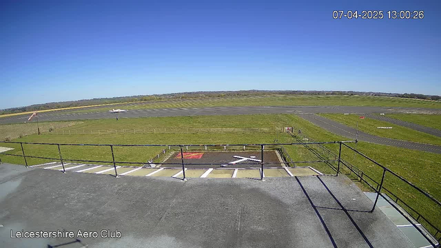 A clear blue sky is visible above an airfield. In the foreground, there is a railing surrounding a viewing area, leading to a tarmac with a prominent white 'X' marking, which indicates a landing pad. A small airplane can be seen taxiing on the runway in the distance. The land around the airfield is green and grassy, with some trees and structures visible in the background.