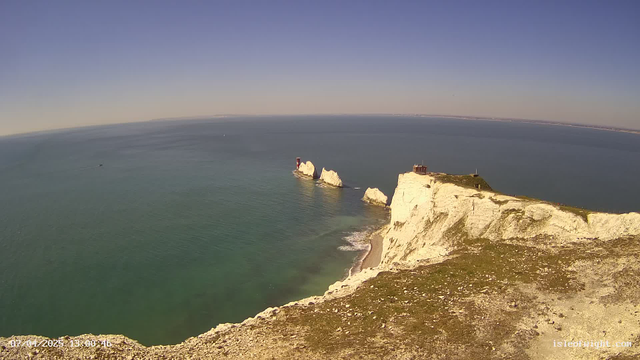 A panoramic view of a coastline featuring white chalk cliffs overlooking a calm, blue sea. In the distance, two prominent chalk stacks emerge from the water, one with a red structure on top. The sky is clear with a gradient of blue and light hues near the horizon. The foreground shows grassy terrain leading to the cliff's edge, with a smooth sandy beach visible below. A small boat can be seen in the water, and the overall scene conveys a sense of tranquility and natural beauty.