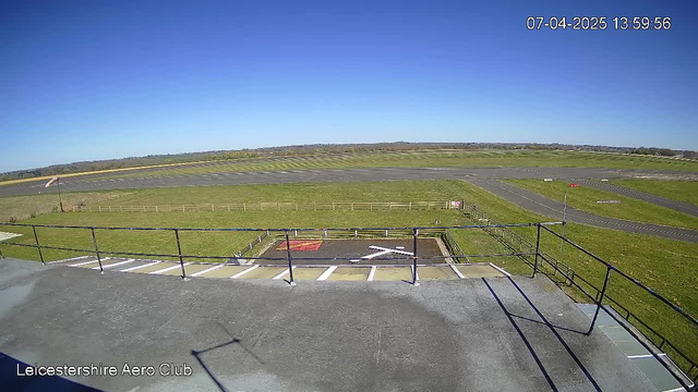 A wide view from a webcam at Leicestershire Aero Club shows a clear blue sky with no clouds. In the foreground, there is a flat rooftop with a railing, and a section of a helipad painted with a large white cross. Below, a grassy area is bordered by a wooden fence. In the background, a runway stretches across the landscape, with another grassy area visible to the left and a hangar in the distance. The scene is bright and sunny, indicating a clear day.