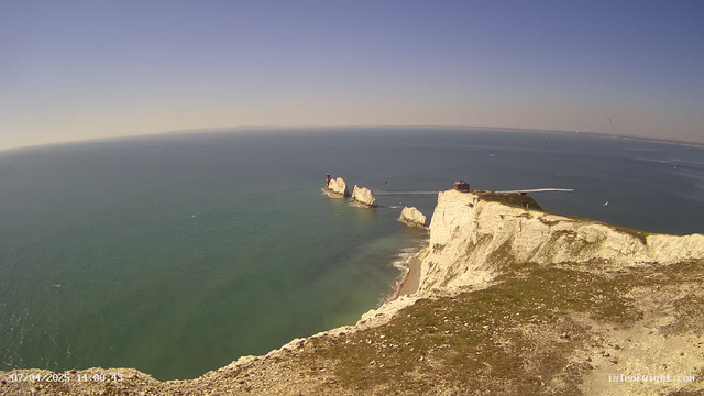 A view of the coastline featuring steep white chalk cliffs rising sharply from the ocean. In the background, the vast blue sea stretches to the horizon under a clear sky. Small boats can be seen on the water, and a few white sails dot the ocean surface. The shoreline has a sandy beach peeking through the rocky terrain, adding texture to the scene.