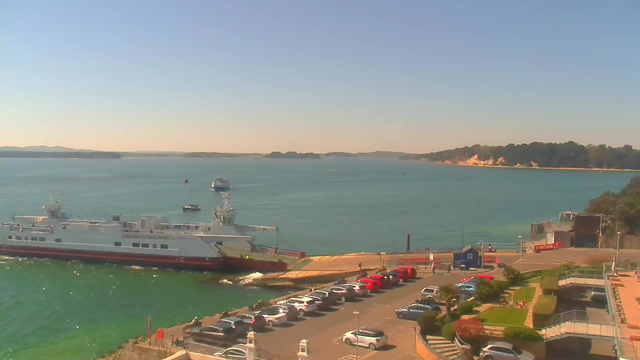 A view of a harbor on a clear day. In the foreground, a large white ferry with red and yellow stripes is docked at the terminal, with some smaller boats visible in the water nearby. A line of parked cars can be seen along the waterfront, with red and white vehicles prominent. The background features calm blue-green water, distant islands, and wooded hills, under a clear blue sky. There are several palm trees and landscaped areas around the terminal building to the right.