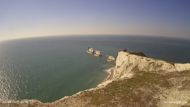 A panoramic view of a coastal area with white chalk cliffs rising steeply above the blue ocean. The clear sky is mostly sunny with a few clouds. In the distance, there are several rock formations emerging from the water. The foreground features grassy areas and rocky terrain along the cliff's edge, and a structure or building can be seen at the top of the cliff. The scene conveys a serene and natural landscape.
