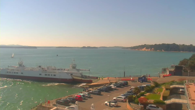 A sunny waterfront scene featuring a large ferry docked at a pier. In the background, there are green hills and a mountainous shoreline. A few small boats, including a sailboat, are on the water. In the foreground, a parking lot is filled with cars, and several people are visible near the ferry. Lush greenery and walkways are also present, adding to the coastal ambiance. The sky is clear and bright.