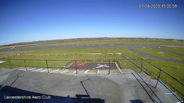 A webcam view of an airfield under a clear blue sky. In the foreground, there is a fenced-off area with a white cross symbol on dark soil. The ground is mostly green grass, and the airstrip in the background has a few visible runway markings. There are also some small flags along the edges of the runway. The overall scene is bright and sunny, with no clouds in the sky.