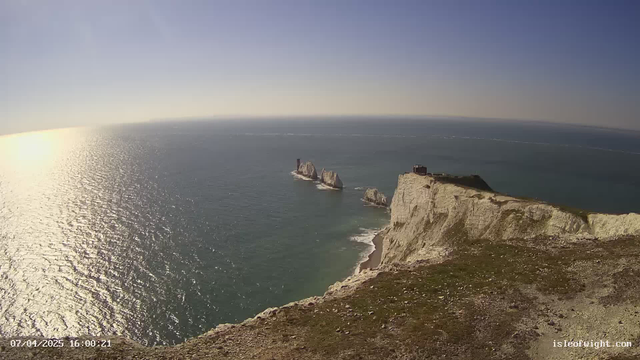 A panoramic view of a coastline featuring white cliffs that drop sharply into the ocean. In the distance, several rock formations rise from the sea. The water appears sparkling and reflective under a clear blue sky. The foreground shows grassy terrain on the cliff's edge, with a building visible atop the cliff near the right side of the image.