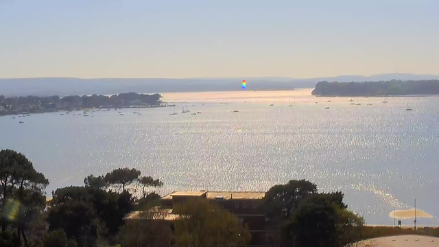 A serene coastal scene showing a body of water reflecting sunlight, creating a shimmering effect. In the distance, several boats are anchored on the water, with gentle hills visible on the horizon. The sky is clear with a light blue hue, and a small rainbow can be seen reflecting off the water's surface. In the foreground, there are trees and a building partially visible, suggesting a lively coastal area.