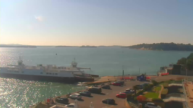 A sunny coastal view showing a docked ferry next to a marina. The calm water reflects the sunlight, and distant hills are visible on the horizon. In the foreground, there is a parking lot with several cars parked, and some greenery is present nearby. The sky is clear with a few wispy clouds.